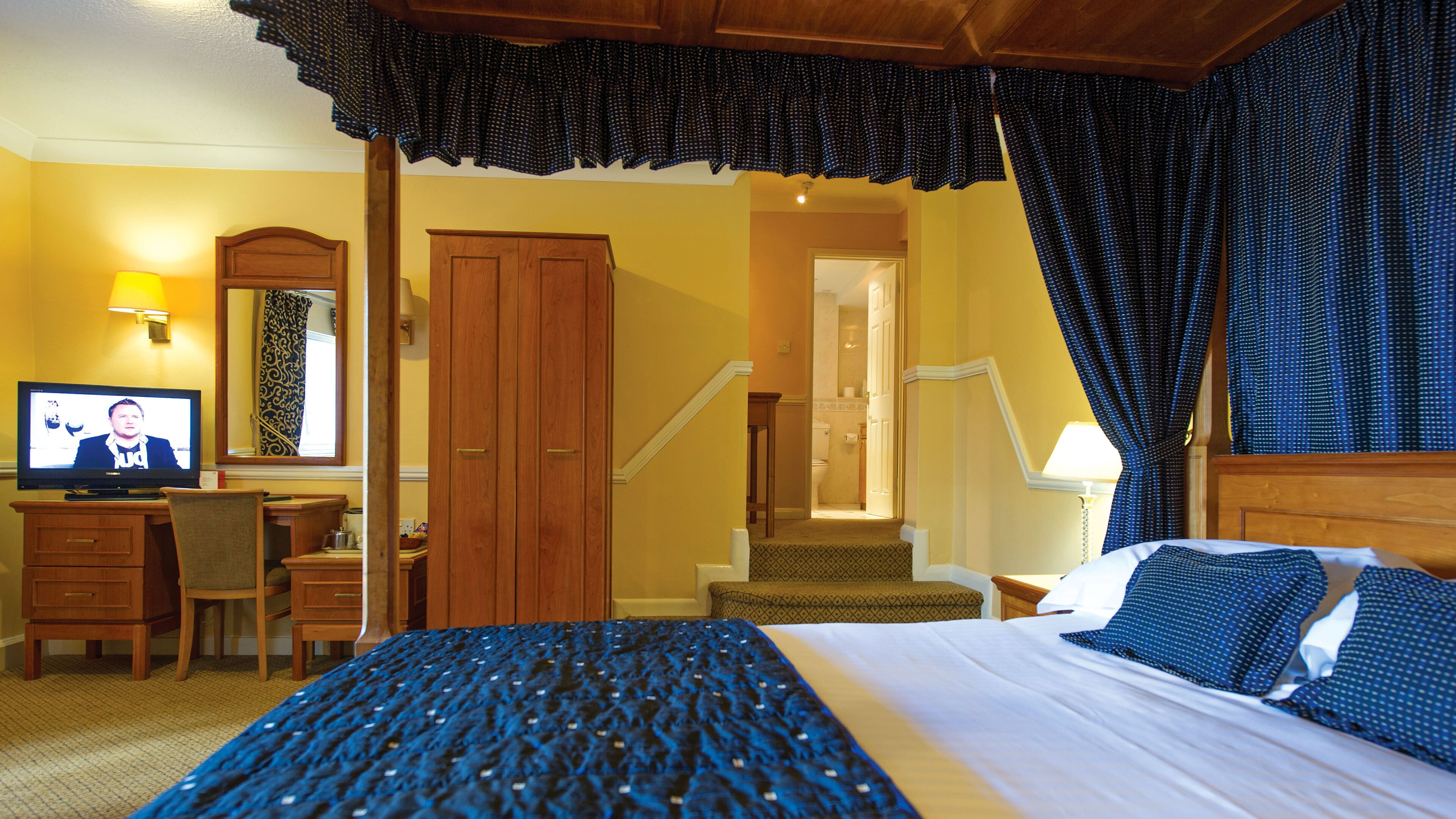 A hotel room with a large bed, a desk with a television, a wooden wardrobe, and a staircase leading to an upper floor.