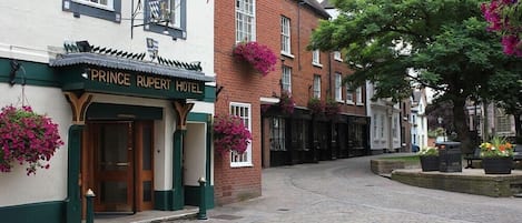 The exterior of the Prince Rupert Hotel with a cobblestone street, hanging flower baskets, and a flag.