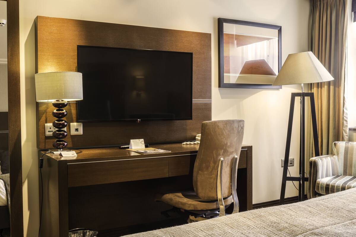 Ein Hotelzimmer mit Flachbildfernseher an einer Holzvertäfelung, einem Schreibtisch mit Lampe, einem gepolsterten Sessel und einem Bett mit gestreifter Decke.