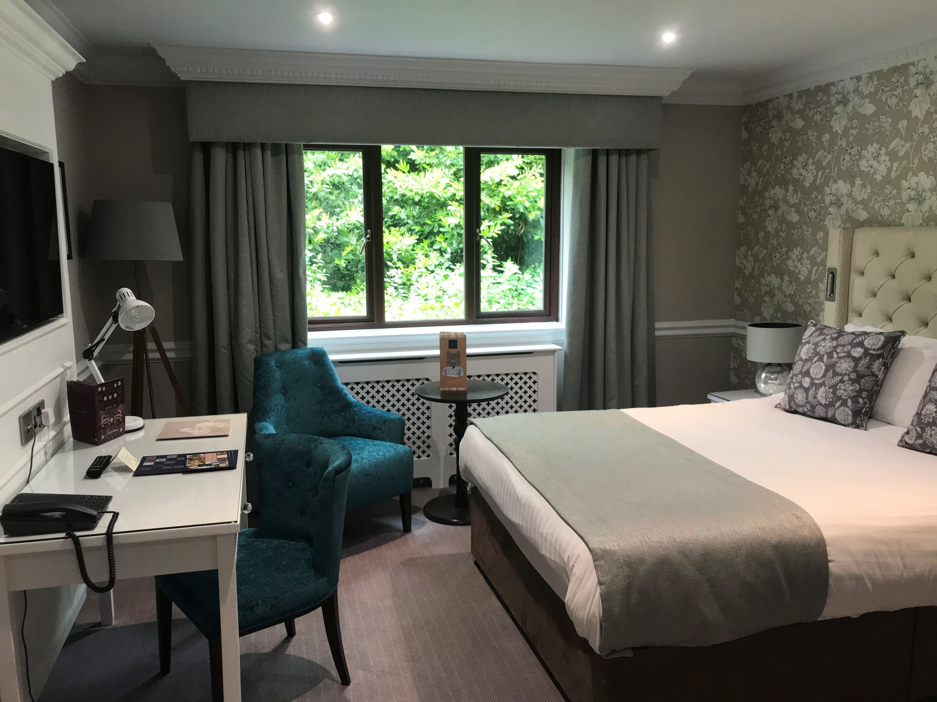 Habitación de hotel con cama, escritorio, silla, ventana con cortinas, lámpara y vistas al exterior con vegetación.