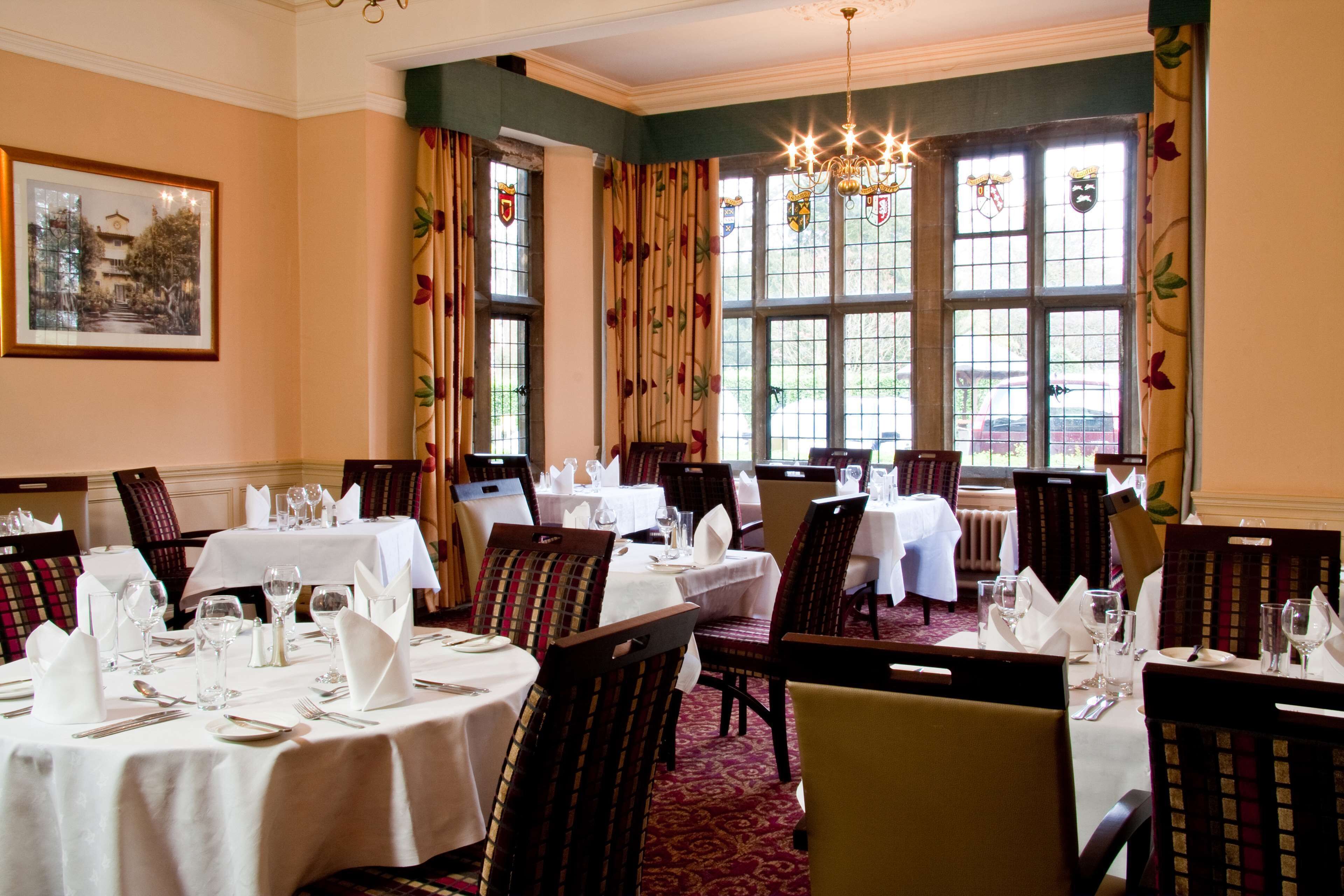 A dining room with multiple tables set for service, large windows with floral curtains, and a chandelier.