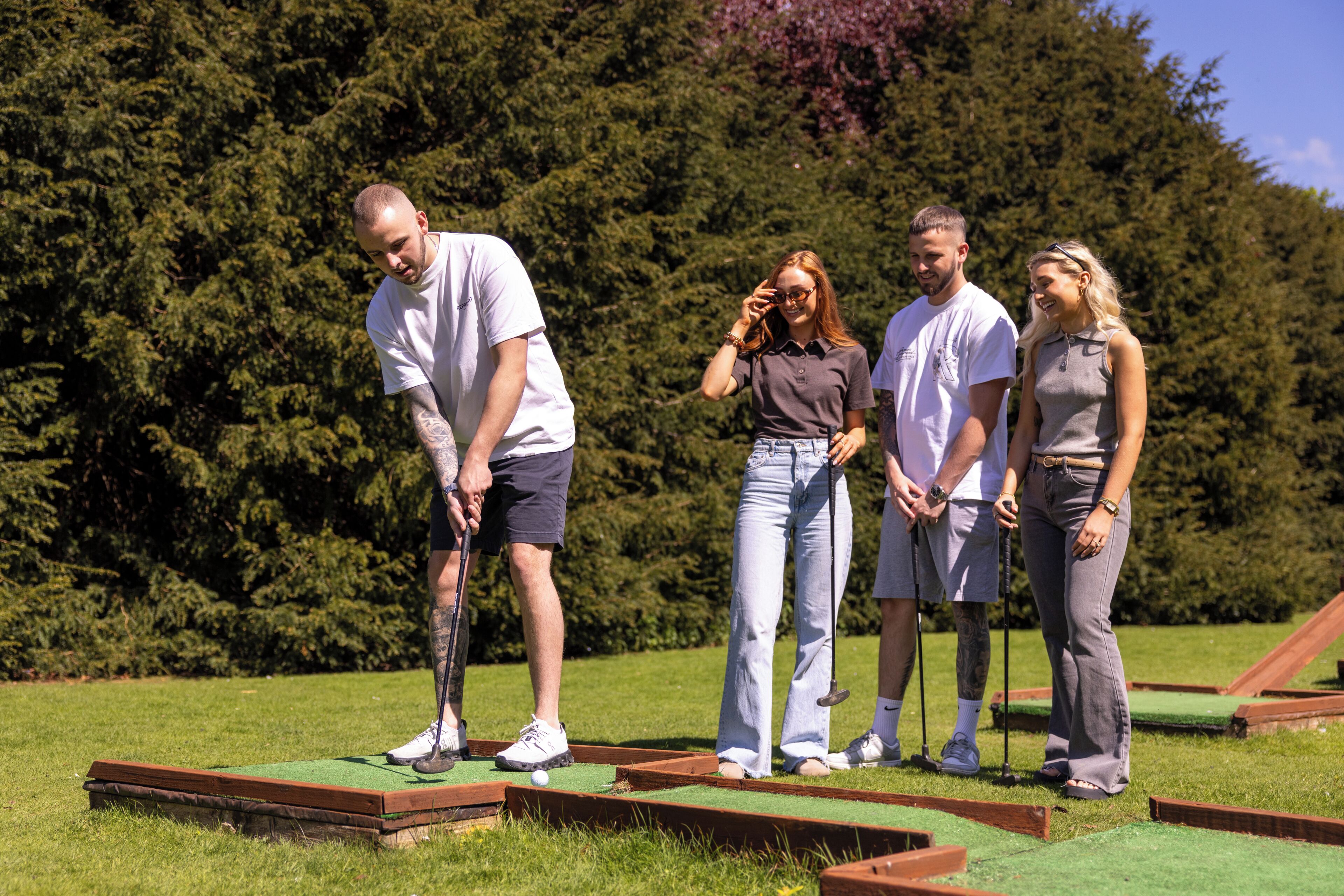 Un grupo de personas jugando minigolf al aire libre, con un campo de césped y árboles de fondo.