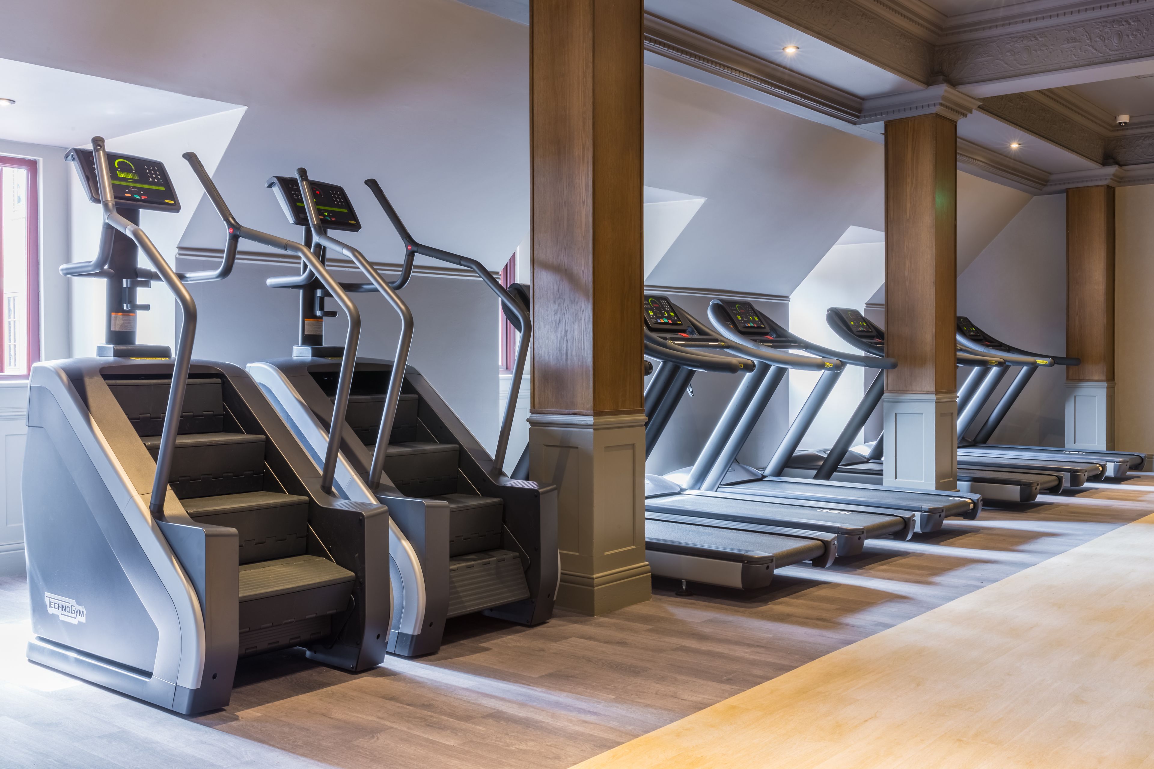 A gym with treadmills and stair climbers, wooden floors, and large windows.