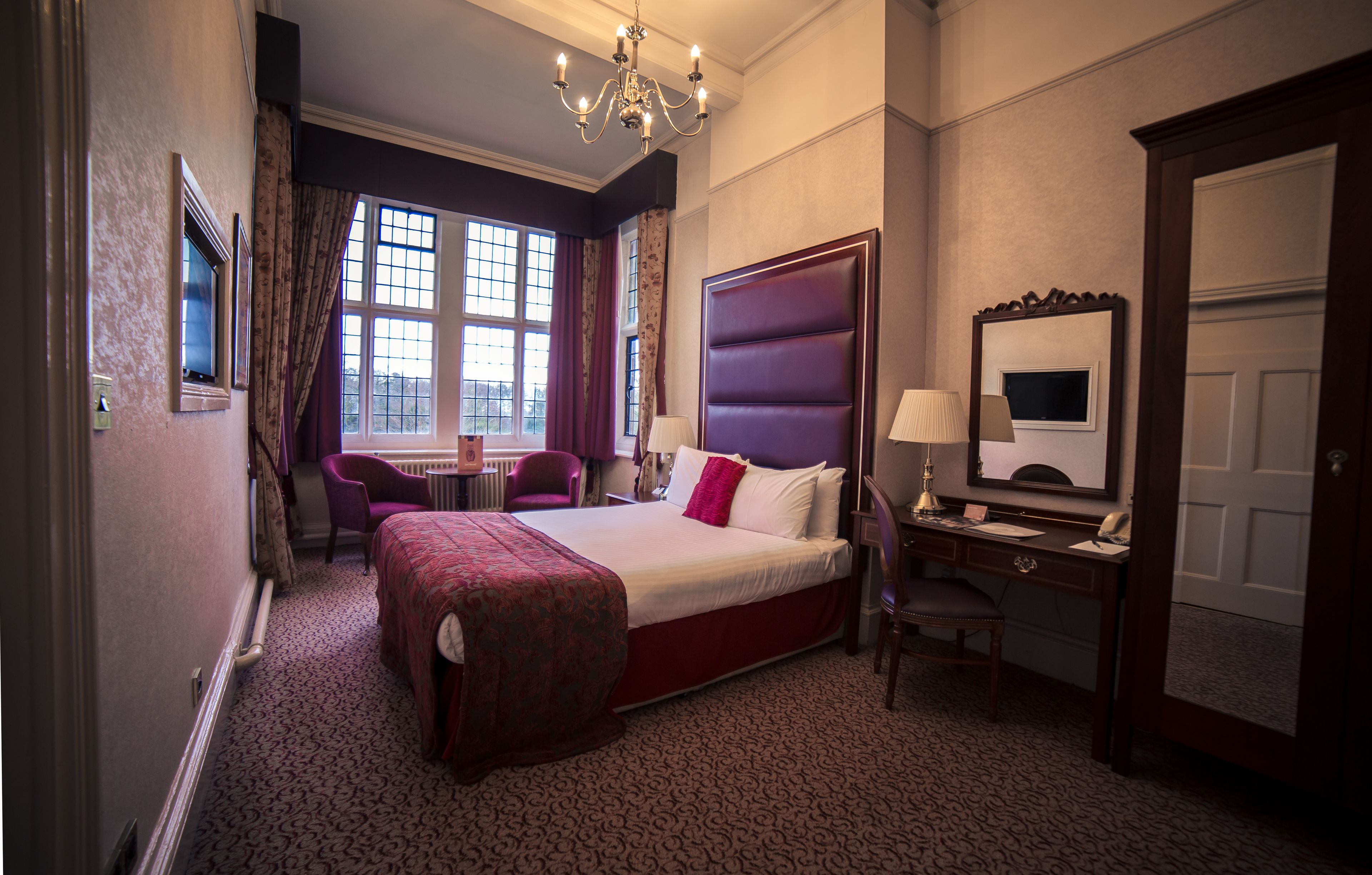 A hotel room with a large bed, a desk with a lamp, a chair, a small table, and a large window with curtains.