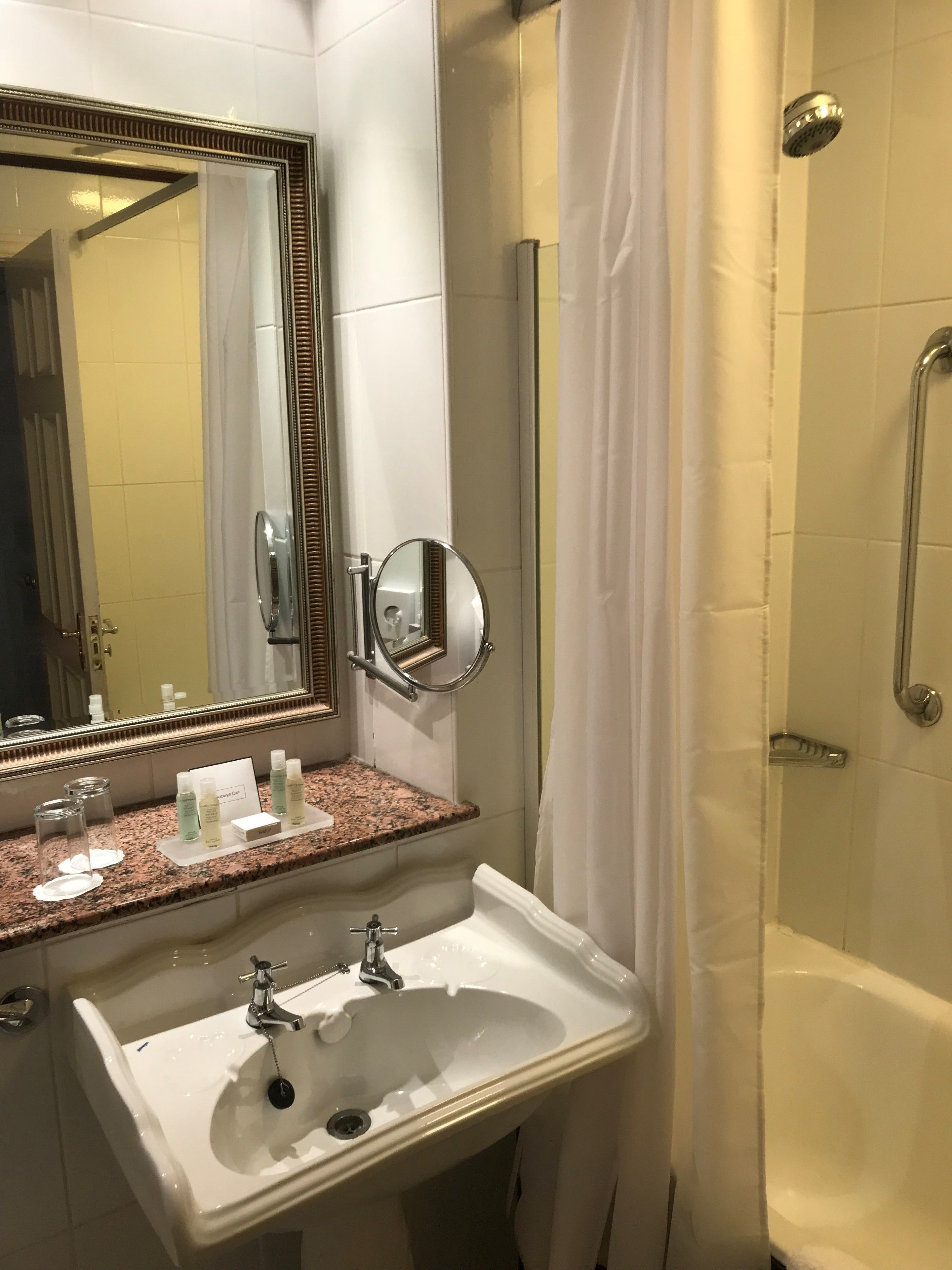 A bathroom with a sink, mirror, shower, and toiletries on the counter.