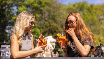Two people toasting with drinks at an outdoor table with a statue in the background.