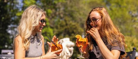 Two people toasting with drinks at an outdoor table with a statue in the background.