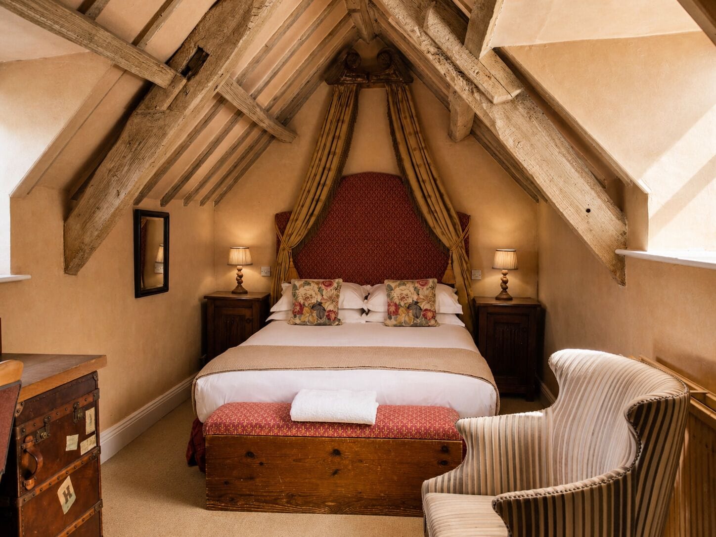 Een slaapkamer met een hemelbed van hout, twee nachtkastjes met lampen, een stoel en een koffer.