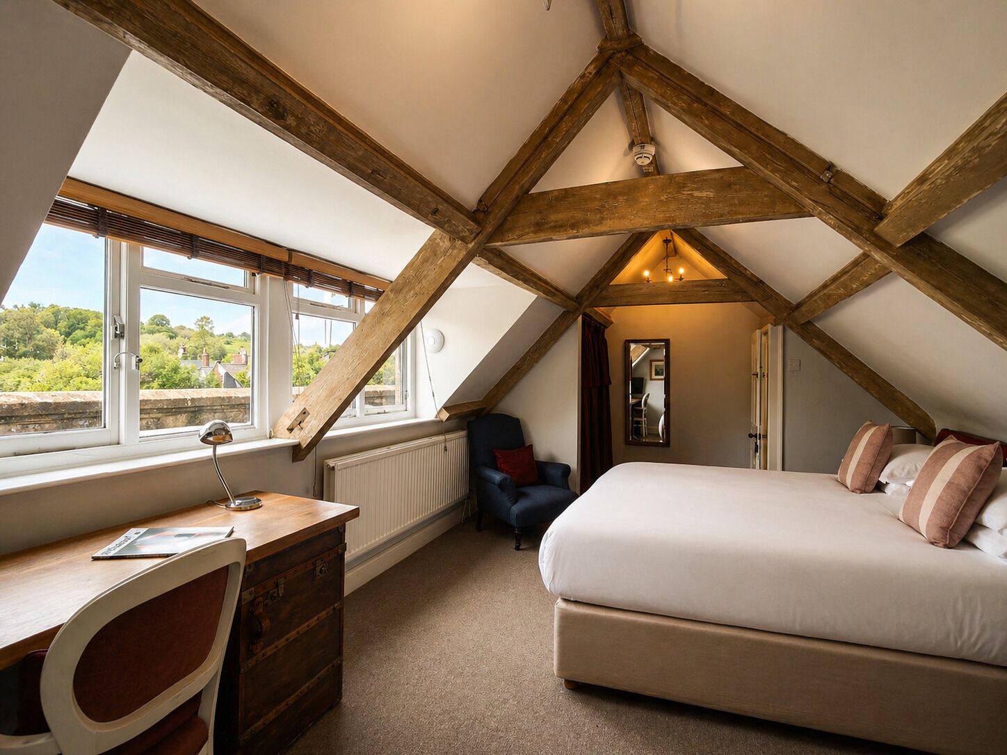 A bedroom with a sloped ceiling, a bed, a desk, a chair, and a window with a view of greenery.