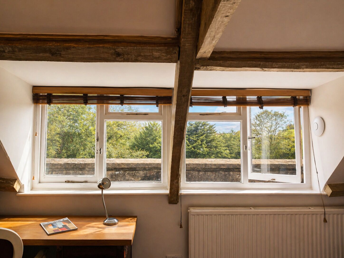 A room with a wooden desk, two windows with blinds, a radiator, and a view of greenery outside.
