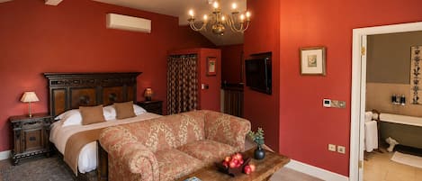 A bedroom with a bed, a sofa, a coffee table with magazines, a chandelier, and a TV.