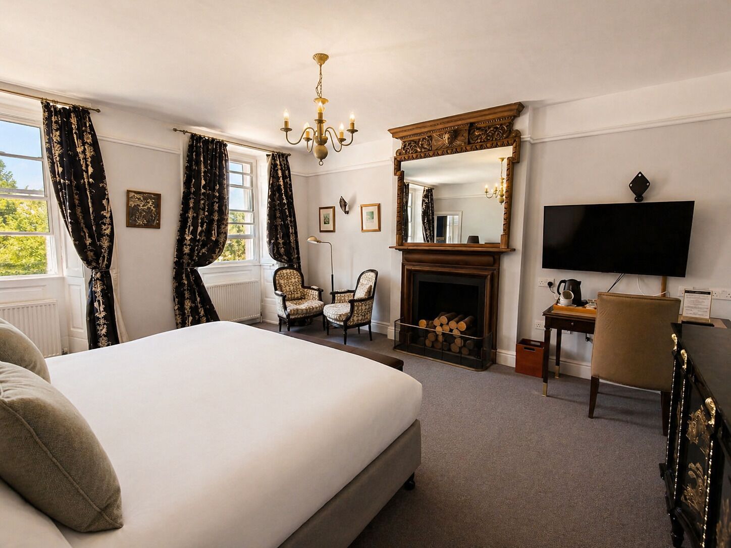 Een hotelkamer met een groot bed, een open haard, een televisie en twee fauteuils.