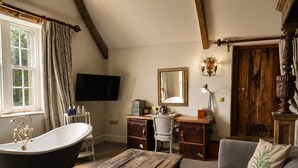 A room with a bathtub, a desk, a chair, a sofa, a television, and a wooden door. - Montigo Resorts, Somerset (Shepton Mallet)