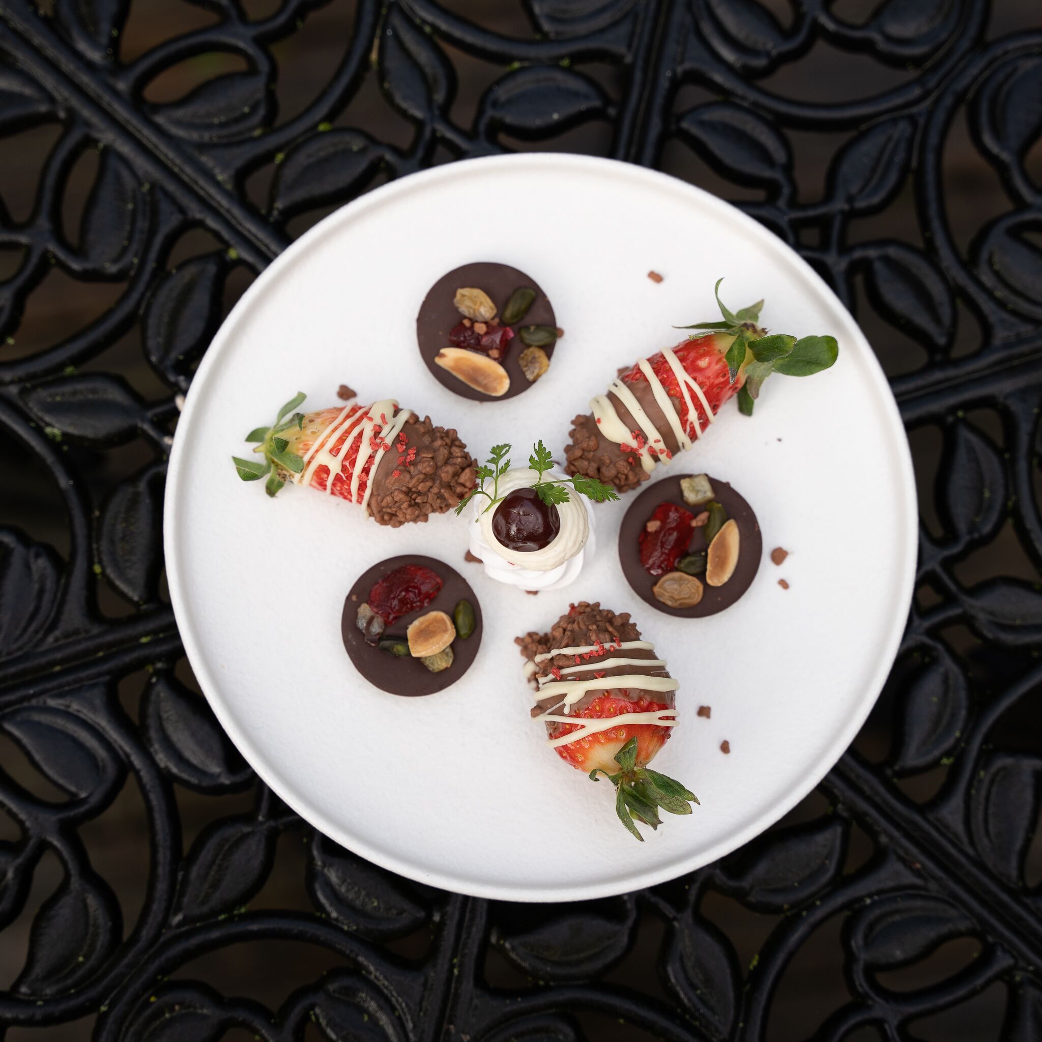 Un plato blanco con fresas bañadas en chocolate y nueces dispuestas sobre una mesa de metal decorativa.