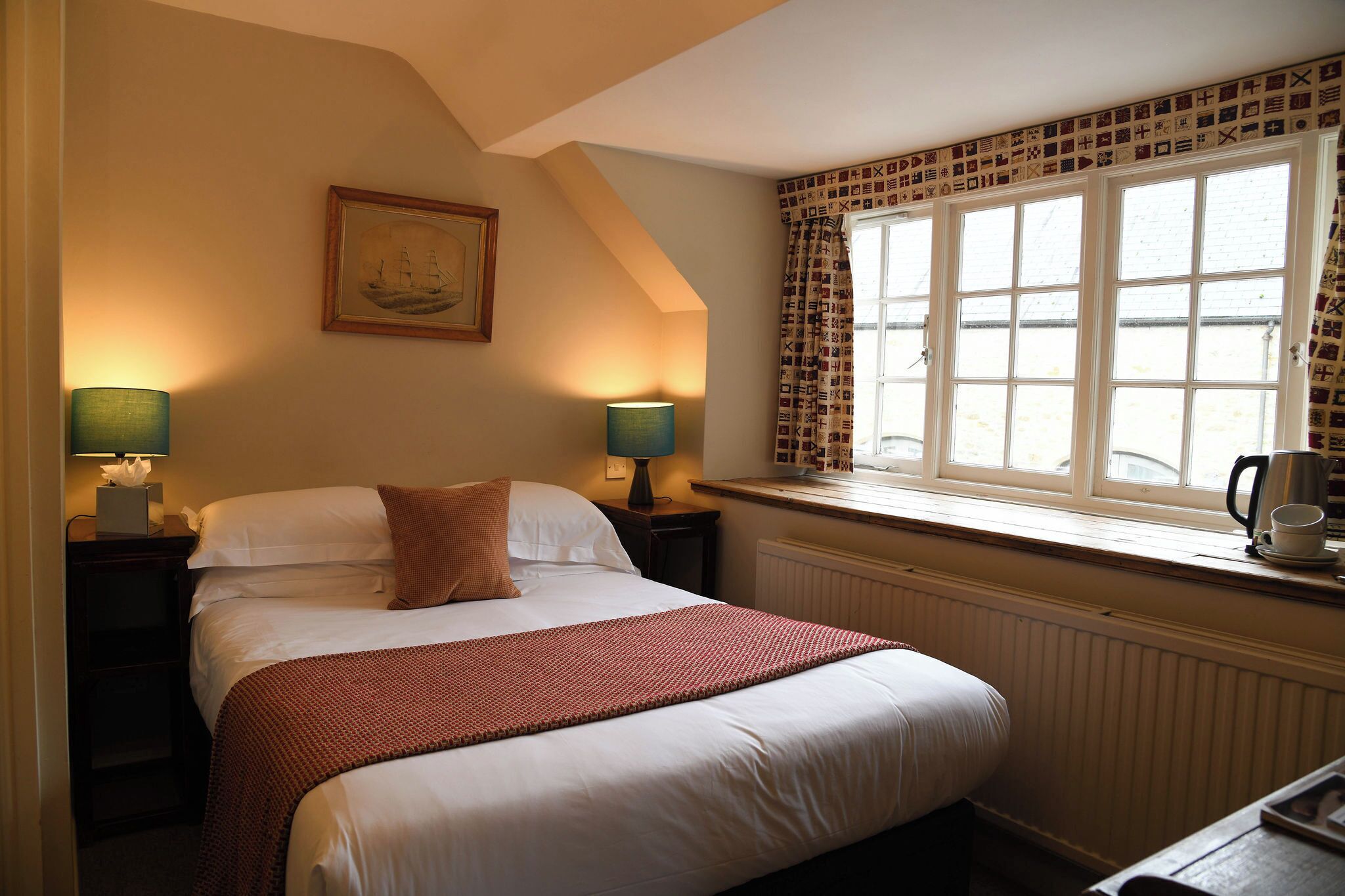 A bedroom with a bed, two bedside lamps, a window with patterned curtains, a radiator, and a framed picture on the wall.