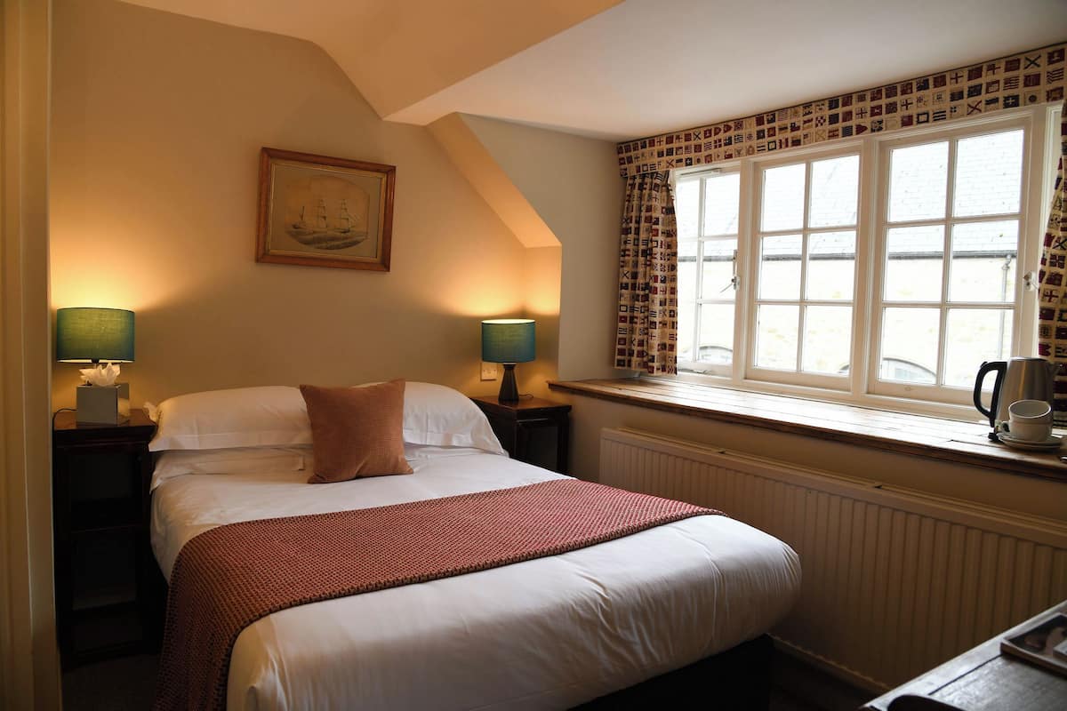 A bedroom with a bed, two bedside lamps, a window with patterned curtains, a radiator, and a framed picture on the wall.