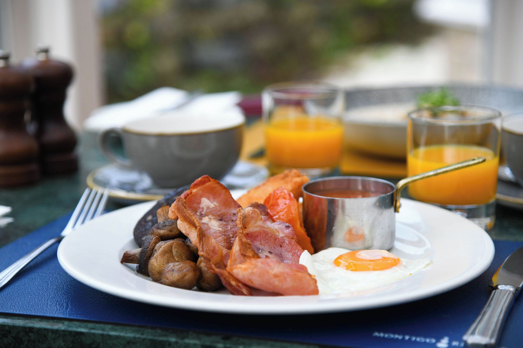 A breakfast plate with bacon, mushrooms, and a fried egg. There are glasses of orange juice and a coffee cup in the background.