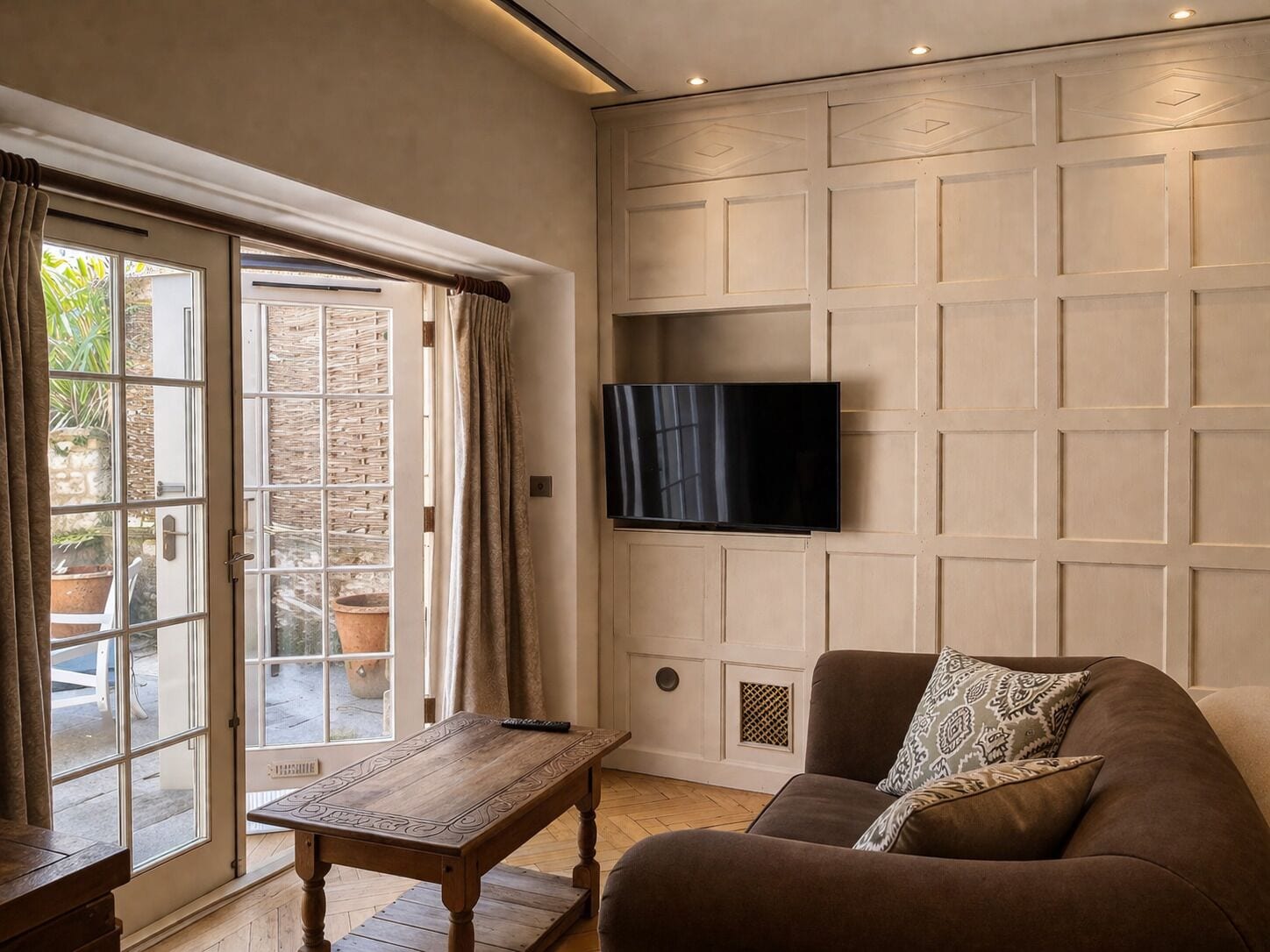 Un salón con televisor de pantalla plana en la pared, un sofá marrón con cojines estampados, una mesa de centro de madera y un ventanal corredero que da a un espacio exterior.