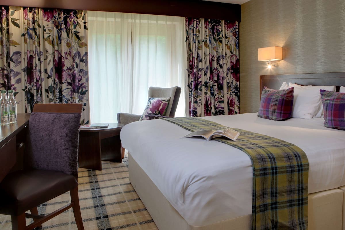 Een hotelkamer met een groot bed, een stoel, een bureau met een lamp en bloemen gordijnen.