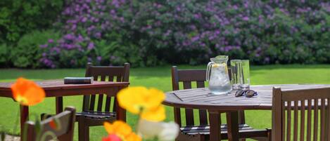 A round wooden table with chairs, a pitcher, and glasses on it, set outdoors with a garden in the background.