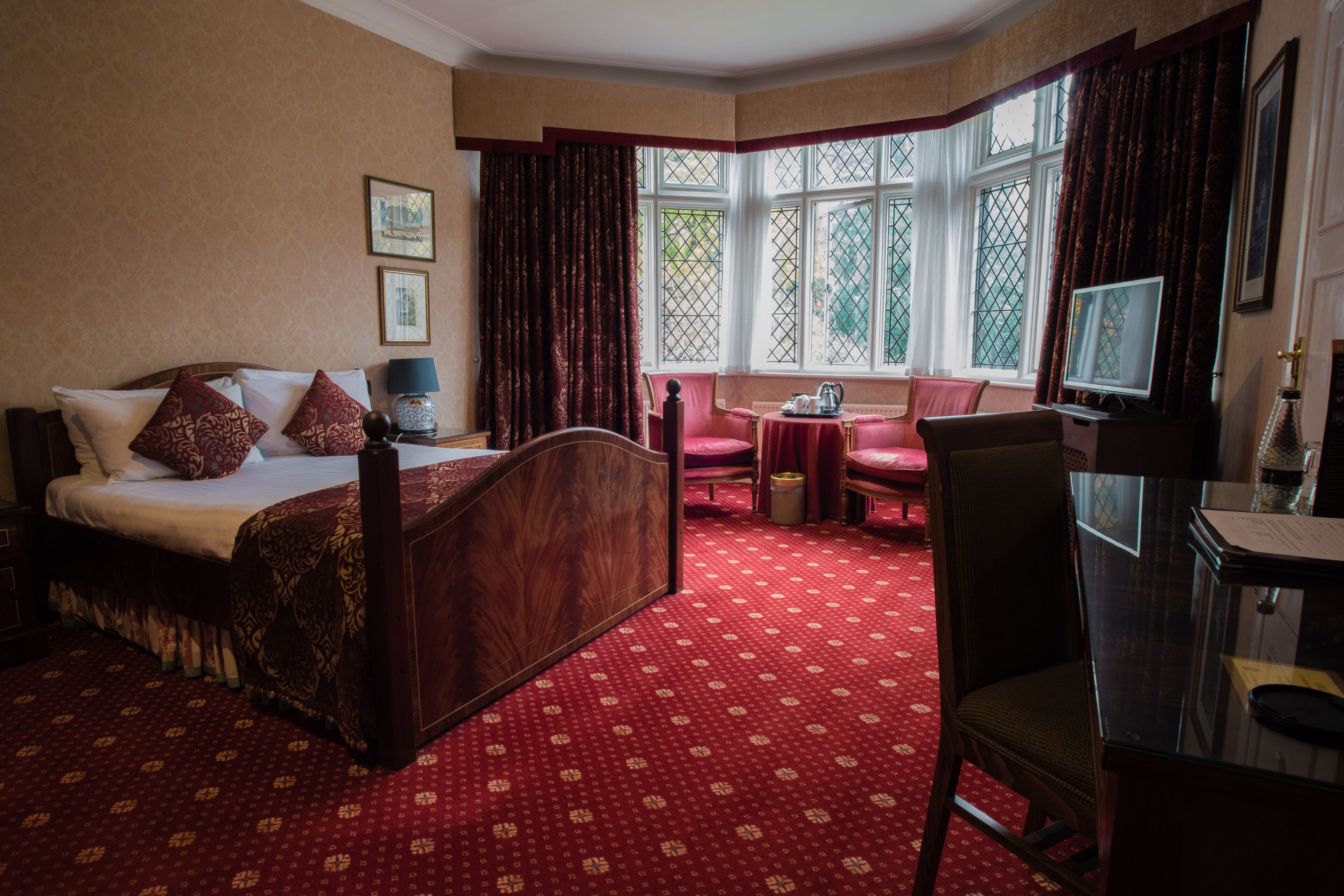 Quarto de hotel com uma cama grande, uma escrivaninha, uma cadeira e uma televisão. Há uma janela com cortinas e vista para o exterior.