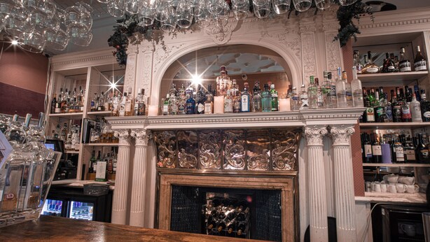 Una zona de bar con chimenea, estantes llenos de botellas de licor y cristalería colgante.