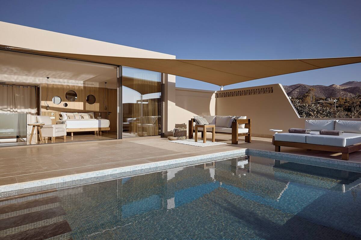 A modern outdoor pool area with a glass-enclosed room, a seating area with a sofa and chairs, and a view of the mountains.
