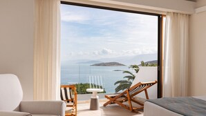 Habitación con un ventanal amplio que ofrece vistas al mar y a las montañas. Se puede ver una cama, una silla y una mesita.