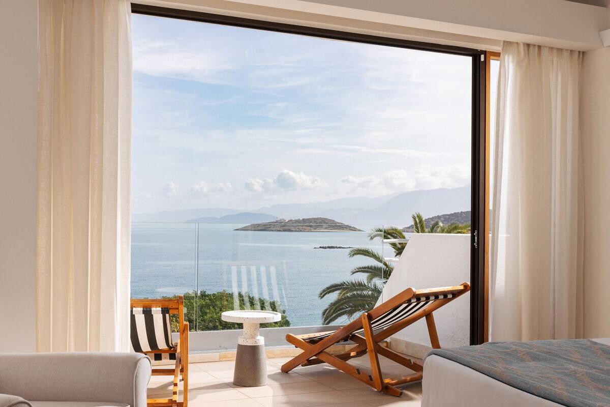 A room with a large window offering a view of the sea and mountains. There is a bed, a chair, and a small table.