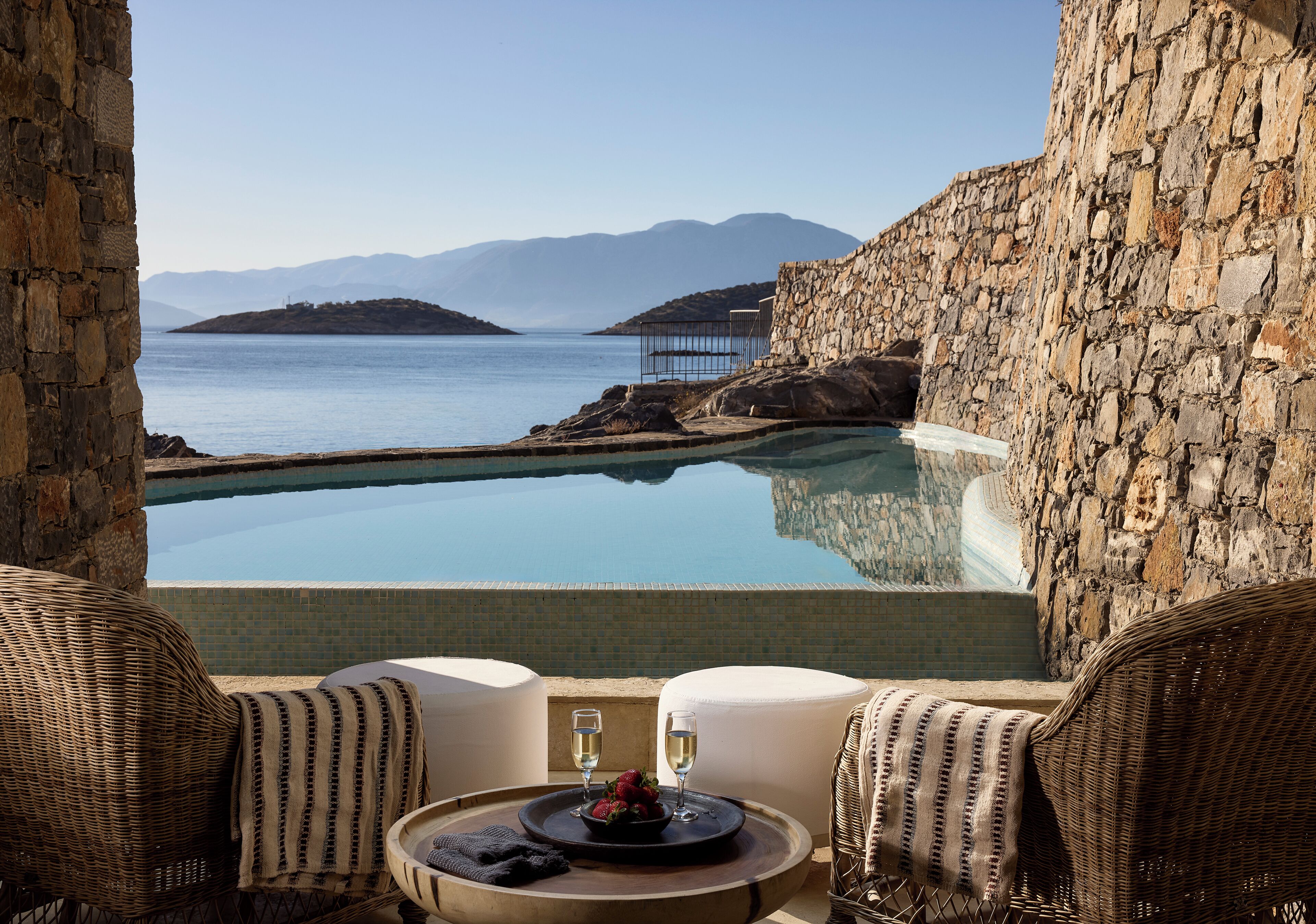 Piscina infinita de piedra con vistas al mar y las montañas, una silla de mimbre y una mesa con una copa de vino y fresas.