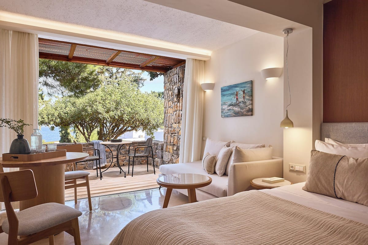 A modern hotel room with a large bed, a sofa, a dining area with a table and chairs, and a view of the outdoors.