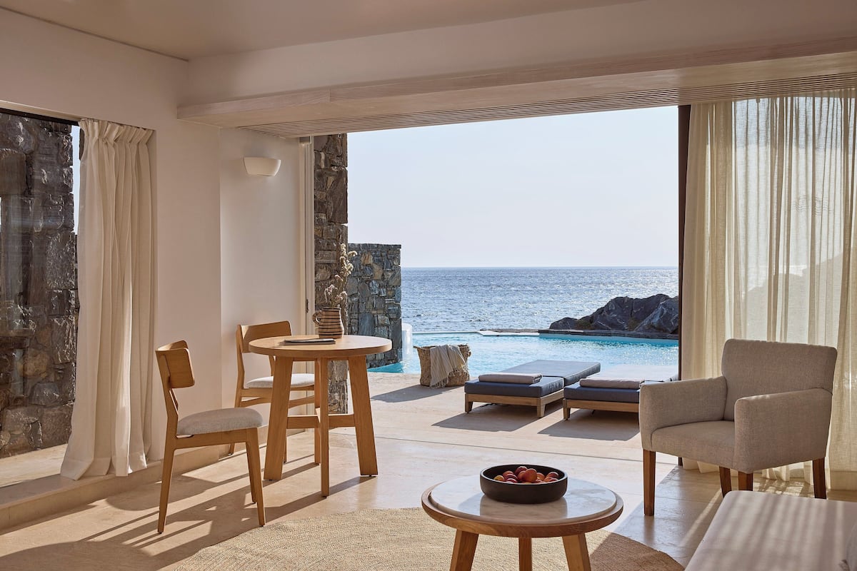 A modern room with a view of the sea, featuring a dining area, a lounge chair, and a table with a bowl of fruit.