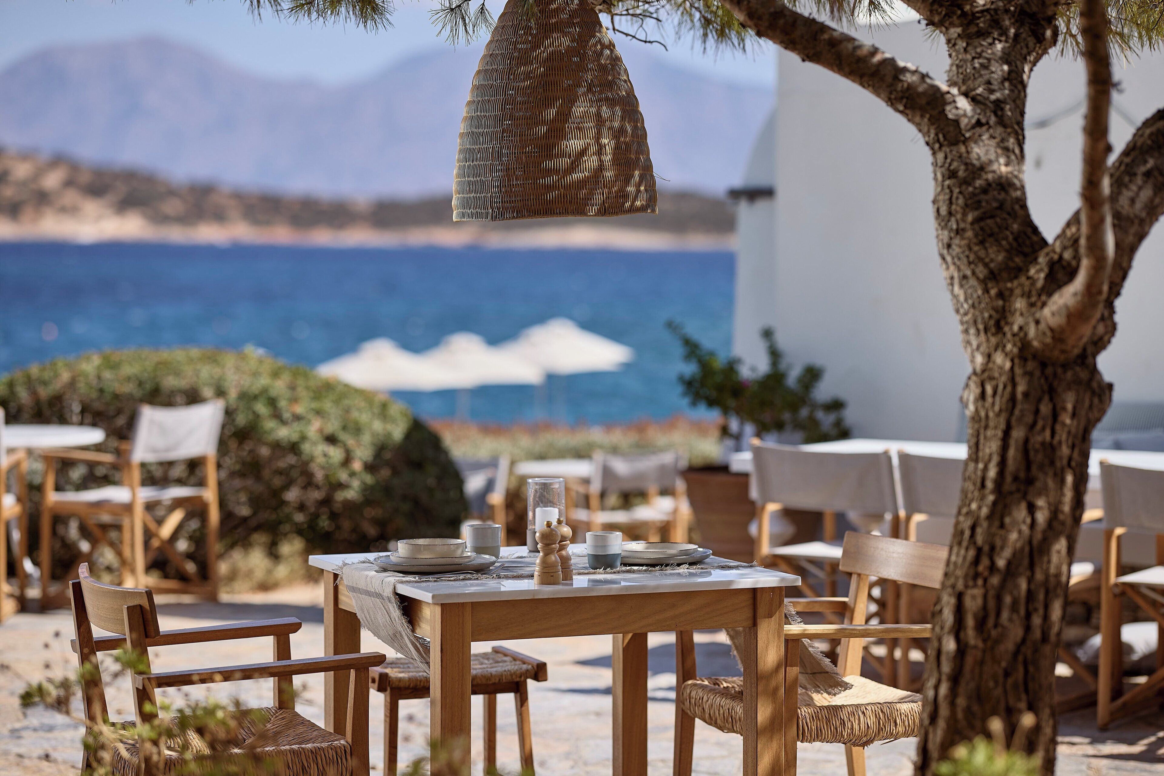 Un área de comedor al aire libre con mesas y sillas de madera, una cesta colgante y vista al mar y montañas en el fondo.