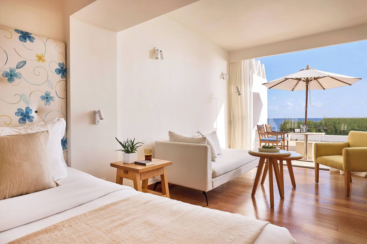 A modern hotel room with a bed, a sofa, a small table with a plant, a chair, and a balcony with an umbrella.