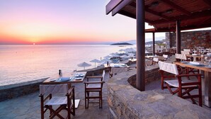 Un área de comedor al aire libre con mesas y sillas, con vista al mar al atardecer.