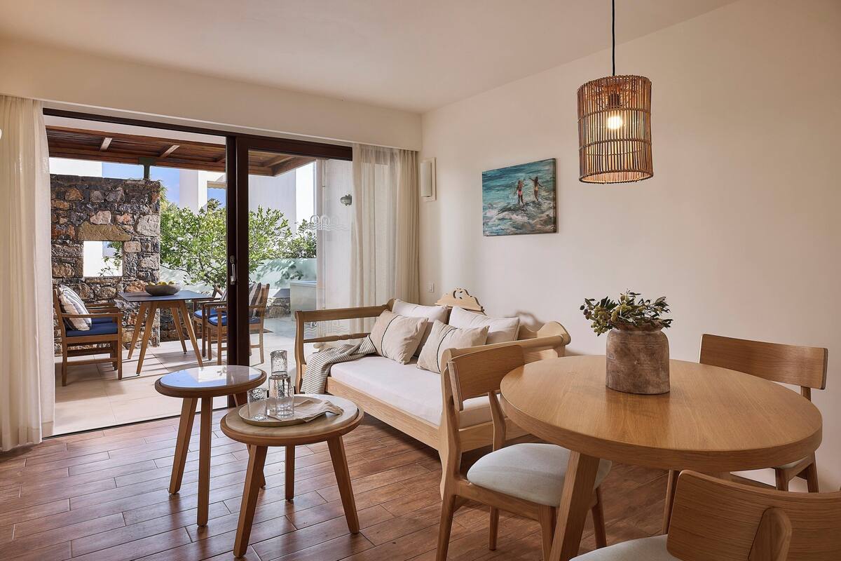 A cozy living area with a wooden sofa, a round wooden table, and a dining table with chairs. There is a stone wall and a view of greenery outside.