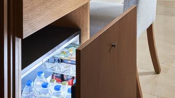 A compact hotel room with a wooden desk, a chair, a mirror, and a mini-fridge stocked with beverages.