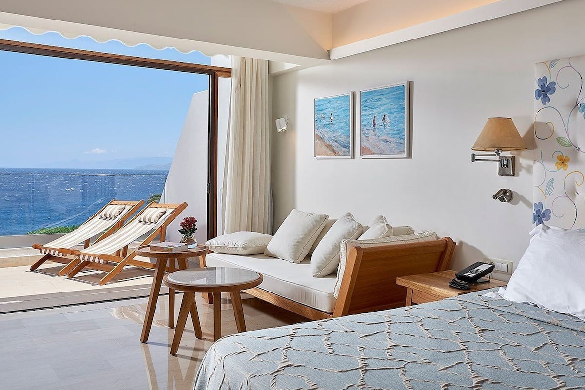 A hotel room with a large bed, a sofa, a small table, and two lounge chairs. There is a view of the ocean through a large window.