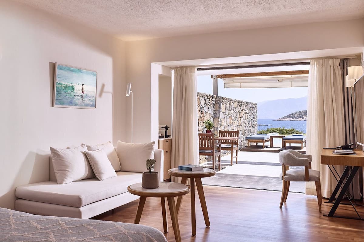 A modern hotel room with a sofa, a small round table, and a view of the sea through an open door.