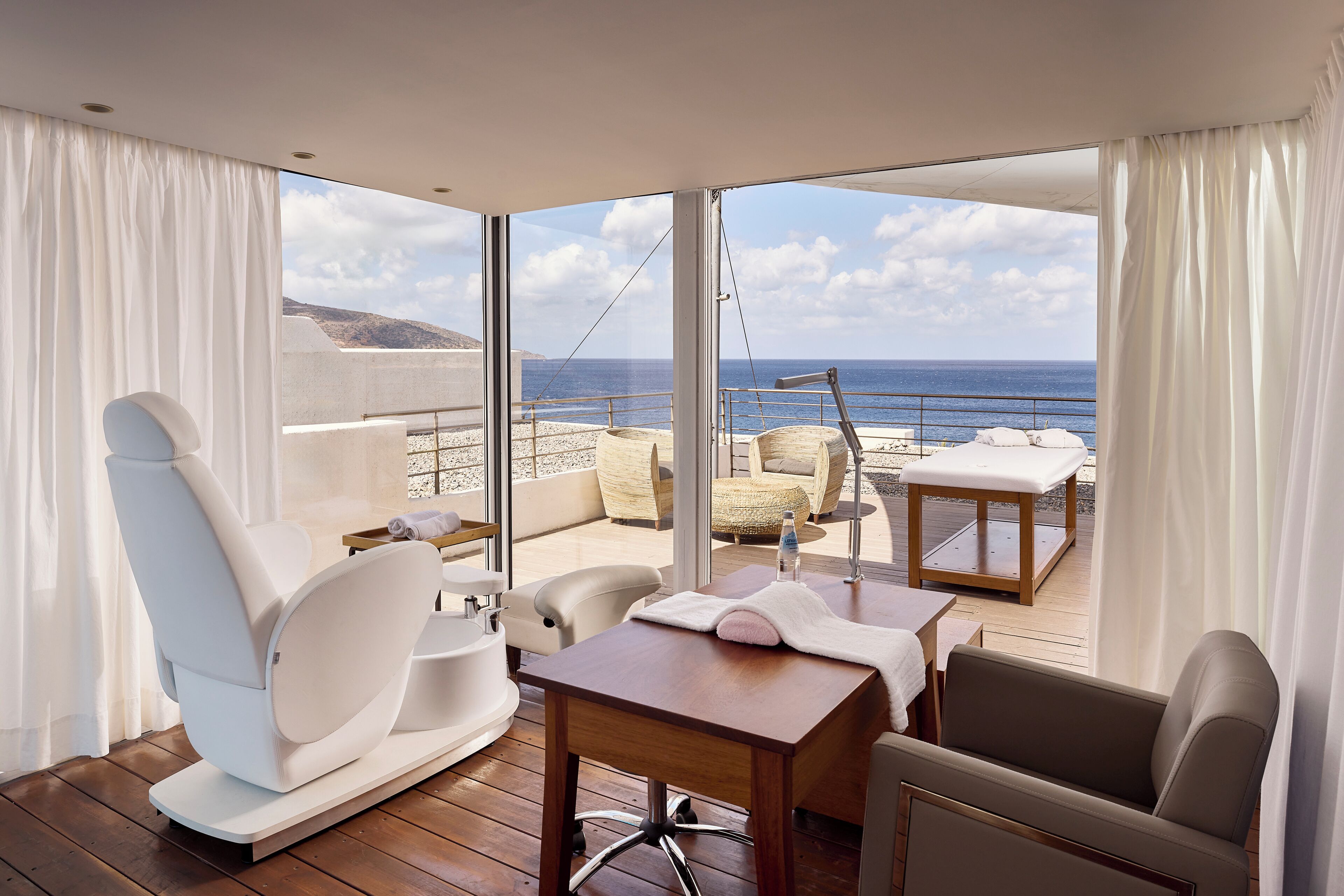 A modern room with a view of the sea, featuring a wooden table, white chairs, a sofa, and a small table with a bottle.