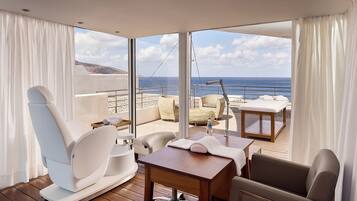 A modern room with a view of the sea, featuring a wooden table, white chairs, a sofa, and a small table with a bottle.