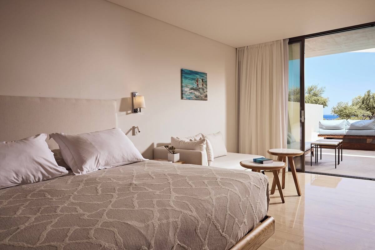 A spacious bedroom with a large bed, a bedside table, a small table, and a view of the outdoors.