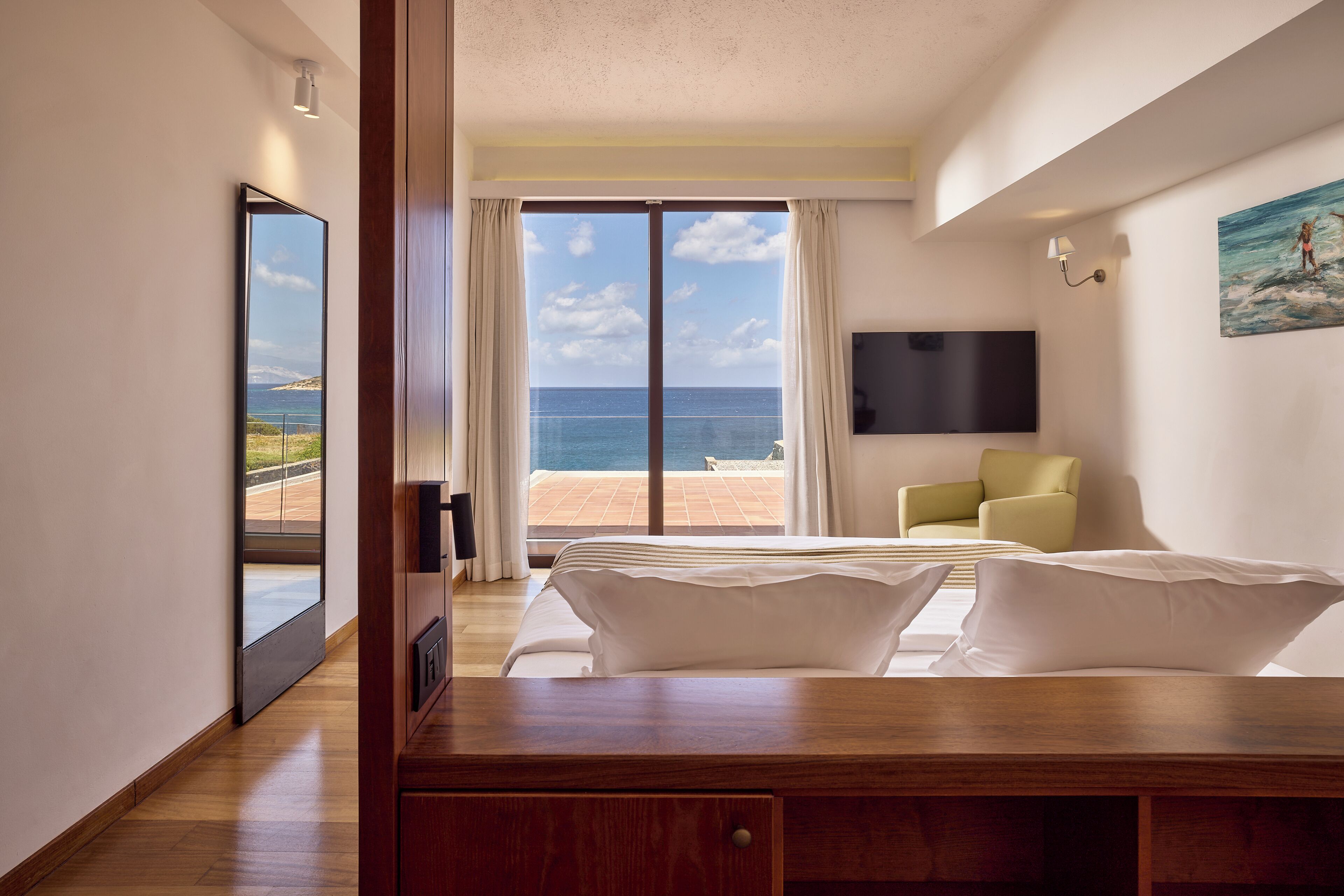 A hotel room with a bed, a TV, a painting, and a view of the sea.