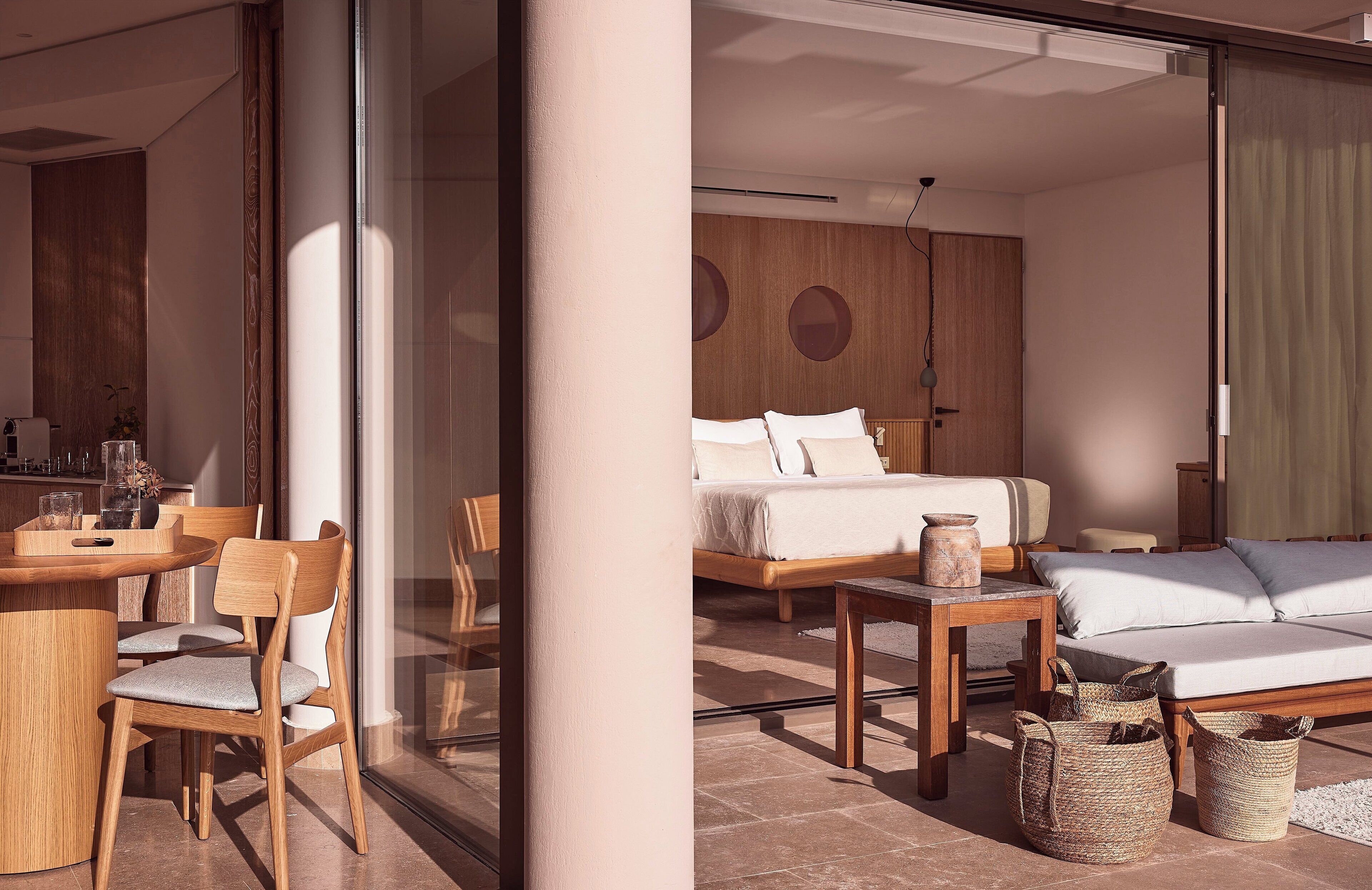 A modern hotel room with a bed, a sofa, a dining table with chairs, and a large window.