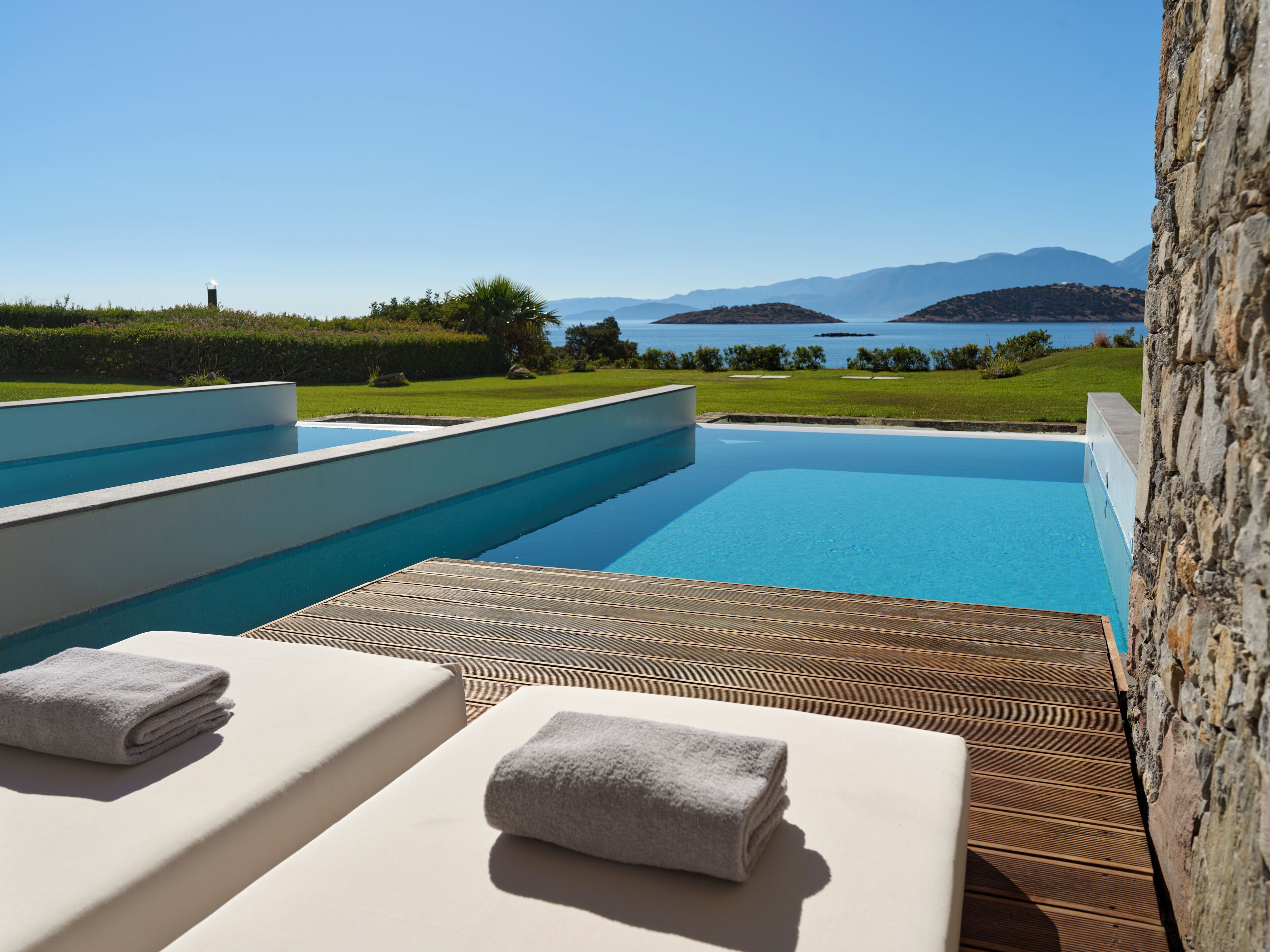 A pool area with two lounge chairs and towels, a wooden deck, and a view of the sea and mountains.