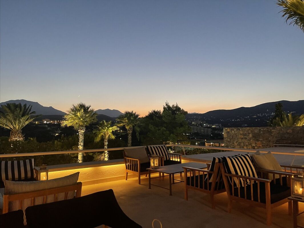 An outdoor seating area with chairs and tables, palm trees, and a view of mountains and city lights at dusk.