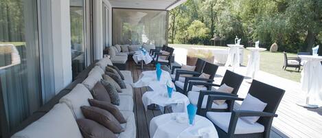 An outdoor seating area with white tables and wicker chairs, a covered patio, and a view of trees and grass.