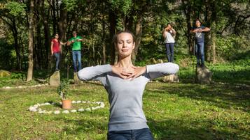 Una persona en una postura de yoga en un parque con árboles y un círculo de piedra al fondo.