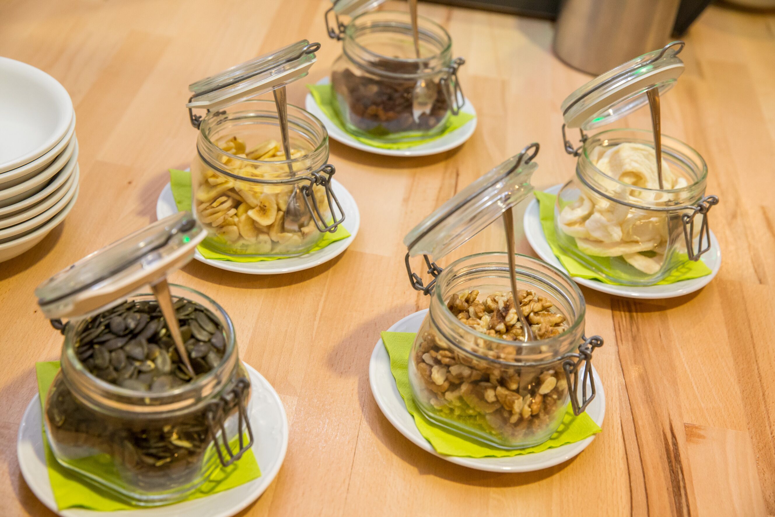 A wooden table with six glass jars containing various nuts and dried fruits, each on a white plate with a green napkin underneath.