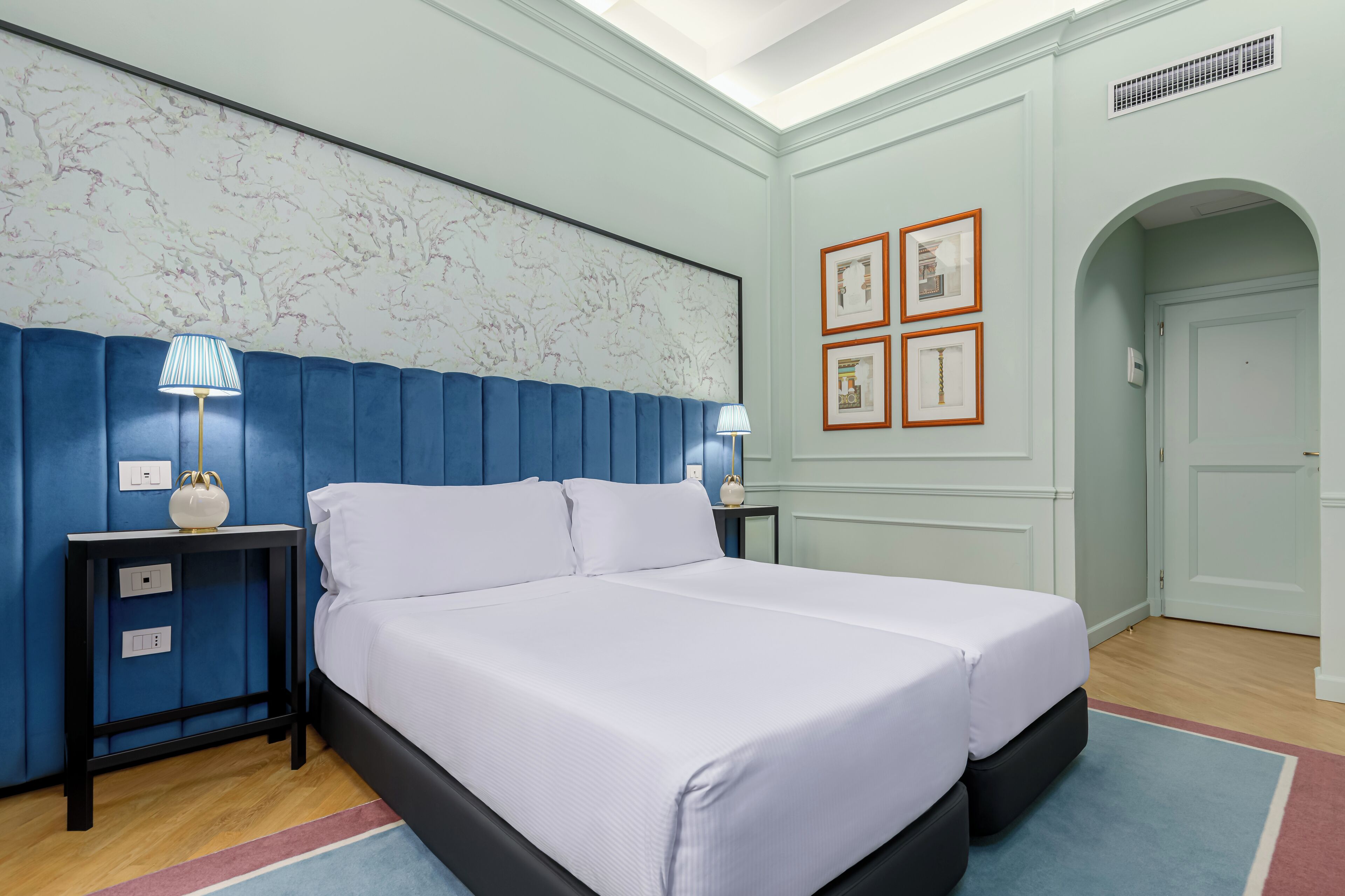 A bedroom with a large bed, two bedside tables, a marble-patterned wall, framed pictures, and a door with an archway.
